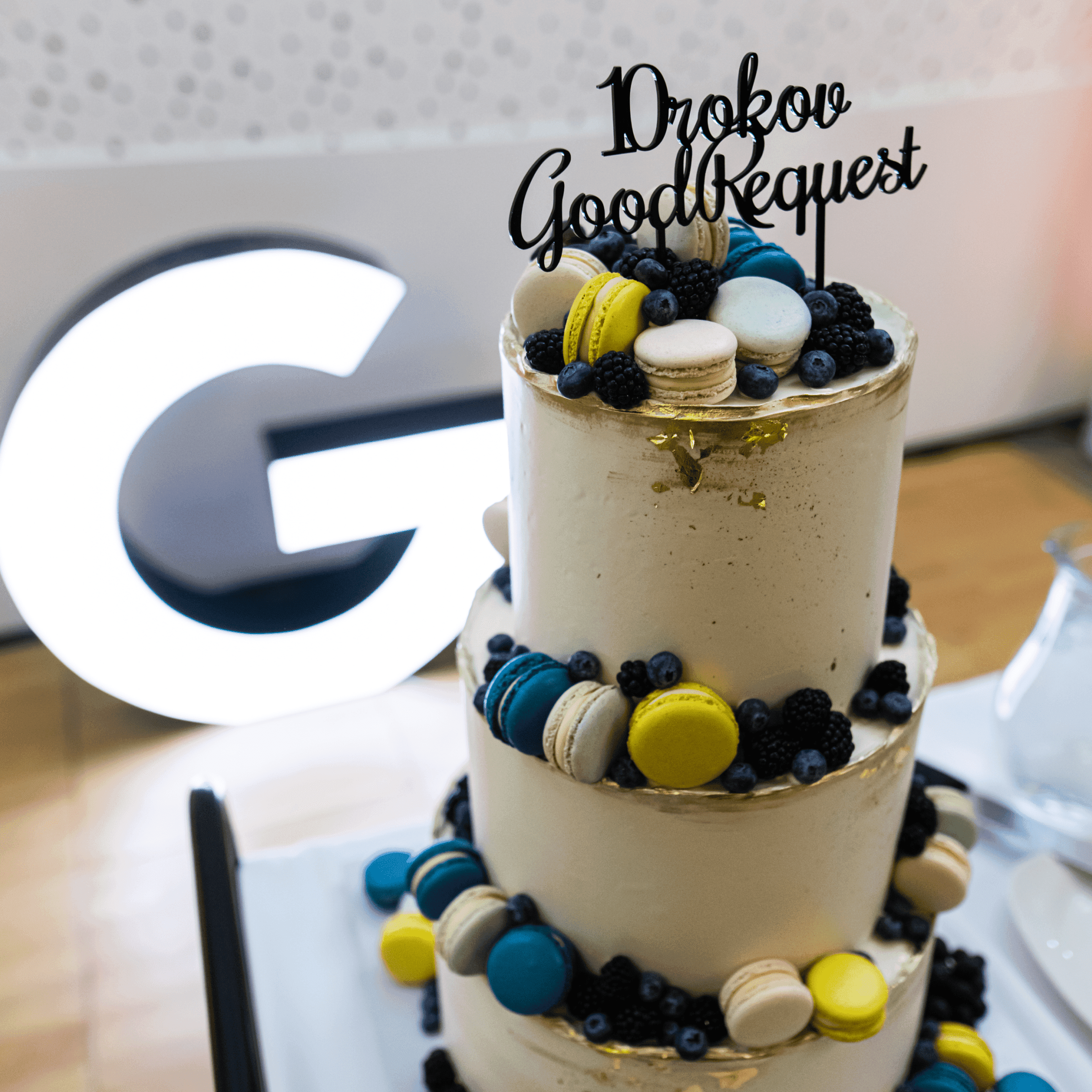 Slávnostná torta s nápisom „10 rokov GoodRequest“, ozdobená makrónkami a lesným ovocím. V pozadí zasvietené logo GoodRequestu.