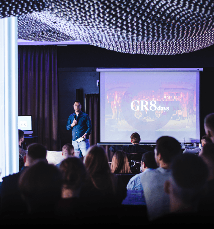 Prezentácia počas podujatia GR8 days, kde pred zaplnenou miestnosťou vystupuje rečník pred veľkou obrazovkou s logom podujatia.