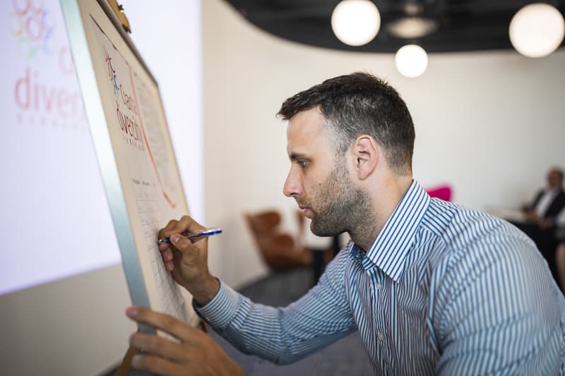 Podpisovanie Charty diverzity zástupcom Goodrequest počas oficiálneho podujatia, s dokumentom umiestneným na stojane v modernom priestore s prítomnosťou ďalších účastníkov.