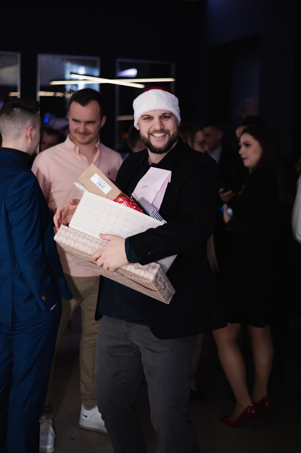 Účastník vianočnej párty Goodrequest s čiapkou Santa Clausa drží niekoľko zabalených darčekov, v pozadí prebieha neformálna konverzácia medzi kolegami v sviatočnej atmosfére.