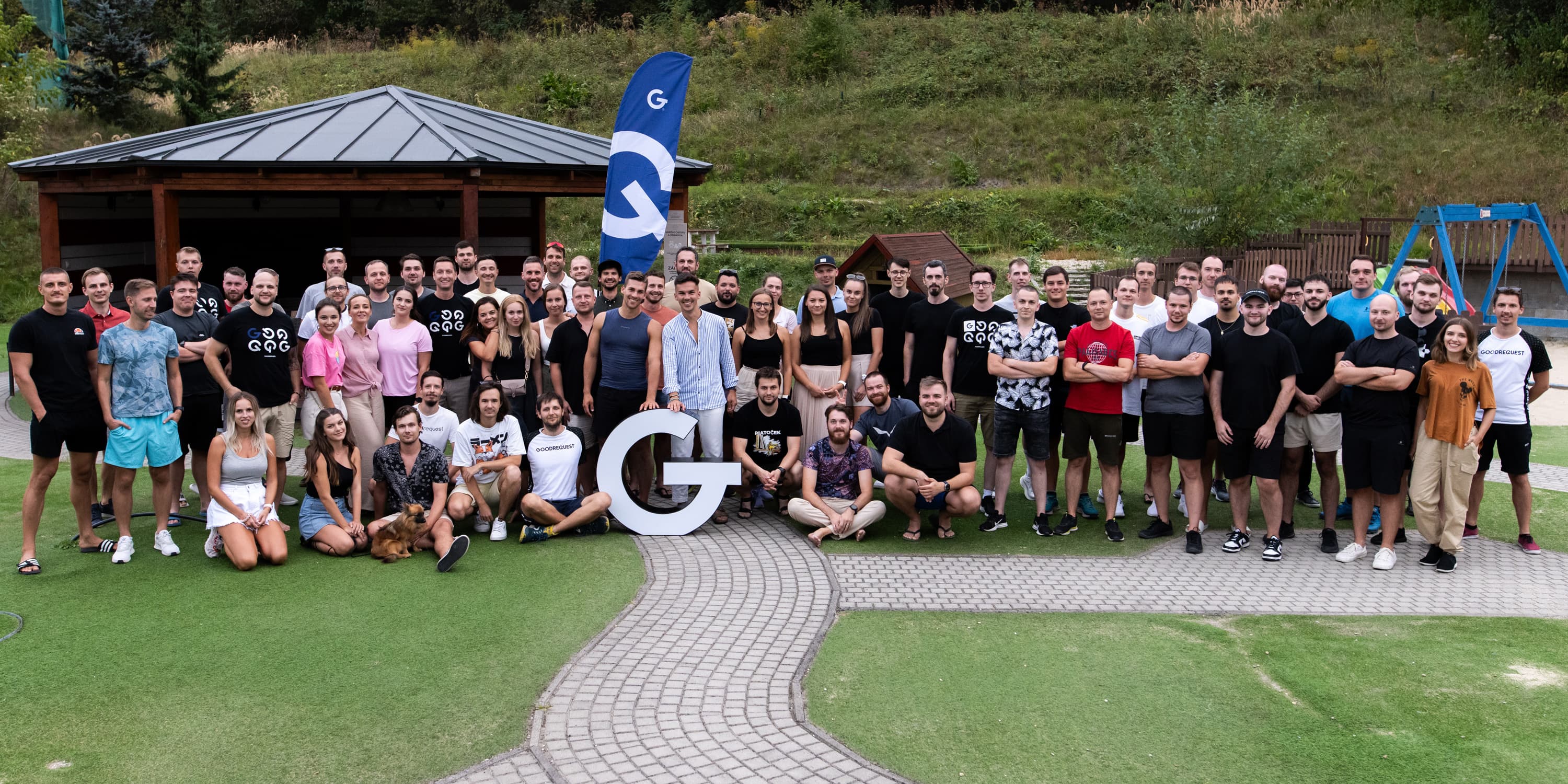 Veľká skupina ľudí pózuje na zelenej tráve počas firemného podujatia pod holým nebom. V popredí je veľké logo GoodRequest, v pozadí sú stromy, altán a modrá obloha.