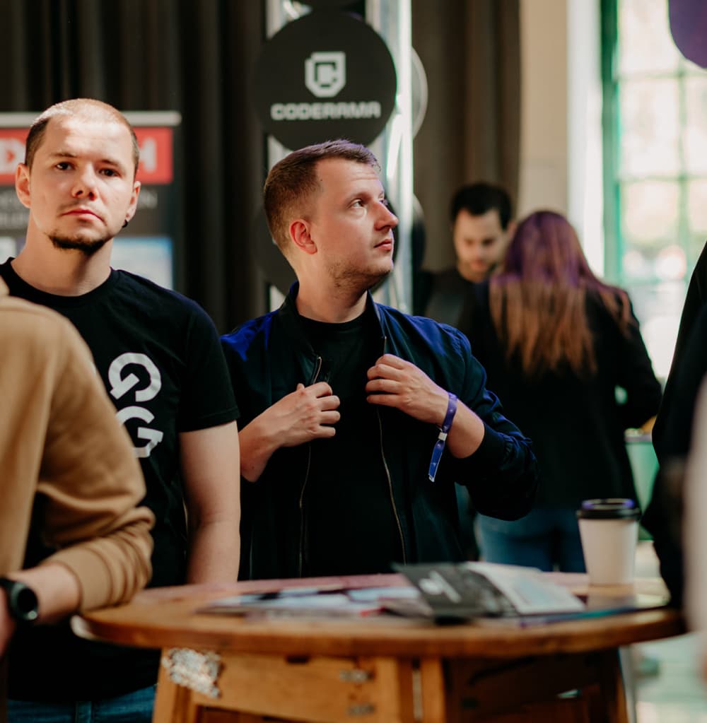 Účastníci technologického podujatia v interiéri, kde dvaja členovia tímu stoja pri stole s dokumentmi a šálkou kávy. V pozadí sú bannery, vrátane loga „CODERAMA“, a ďalší ľudia zapojení do neformálnych rozhovorov.