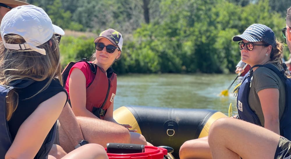 Skupina členov tímu Goodrequest počas splavu, sediaca v nafukovacom člne na pokojnej rieke, obklopená zeleňou. Všetci majú záchranné vesty a pokrývky hlavy, zachytení v uvoľnenej atmosfére teambuildingovej aktivity v prírode.