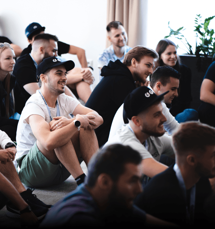 Členovia tímu sediaci na koberci, počas zahájania jedn=ho z teambuildingov spoločnosti GoodRequest. Účastníci sa usmievajú a sú sústredení na dianie pred nimi.