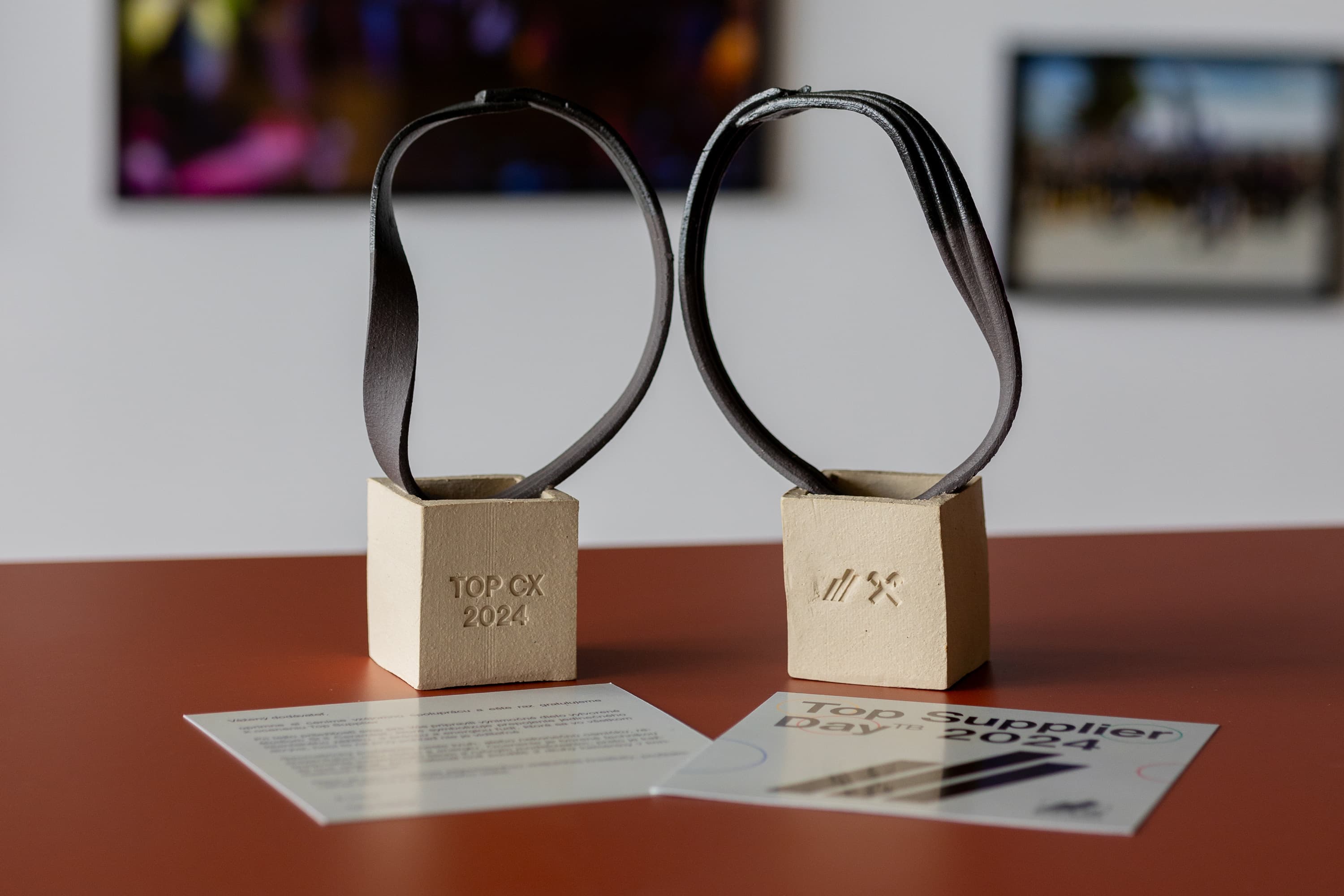 Dva ocenenia na stole s kovovou štruktúrou. Na jednom je nápis "TOP CX 2024", pred nimi sú dve karty. V pozadí sú rozmazané fotografie na stene.