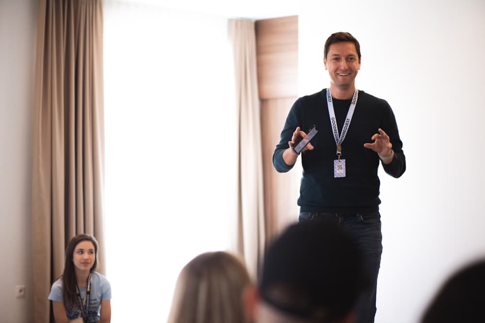 Člen vedenia Goodrequest stojí pred tímom počas teambuildingového podujatia v interiéri, s identifikačnou kartou na lanku a prezentačným ovládačom v ruke. V pozadí sú viditeľné závesy a okno.