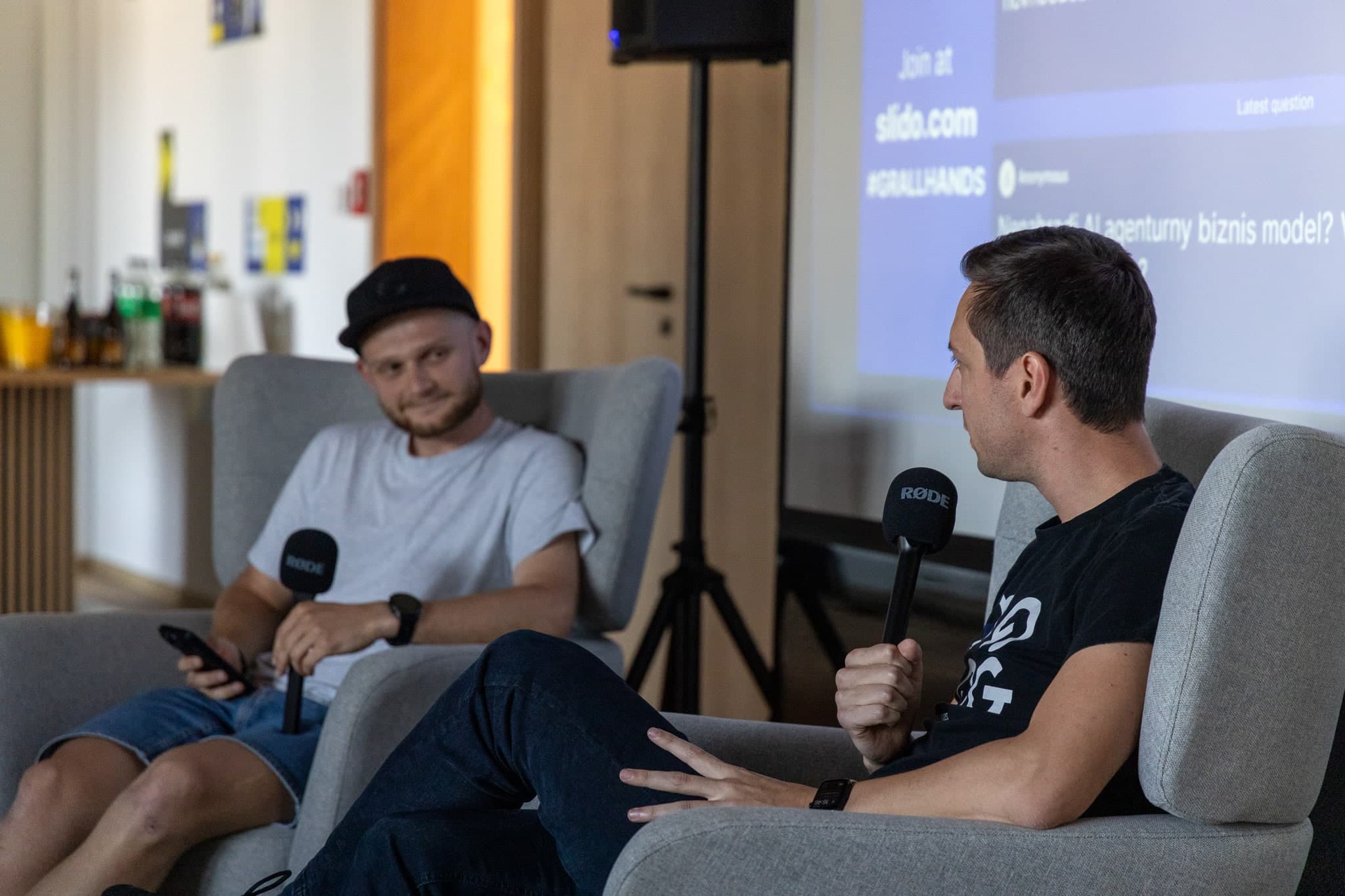 Fotografia dvoch mužov s mikrofónmi pred veľkým plátnom a reproduktorom v pozadí, jeden z nich práve rozpráva.