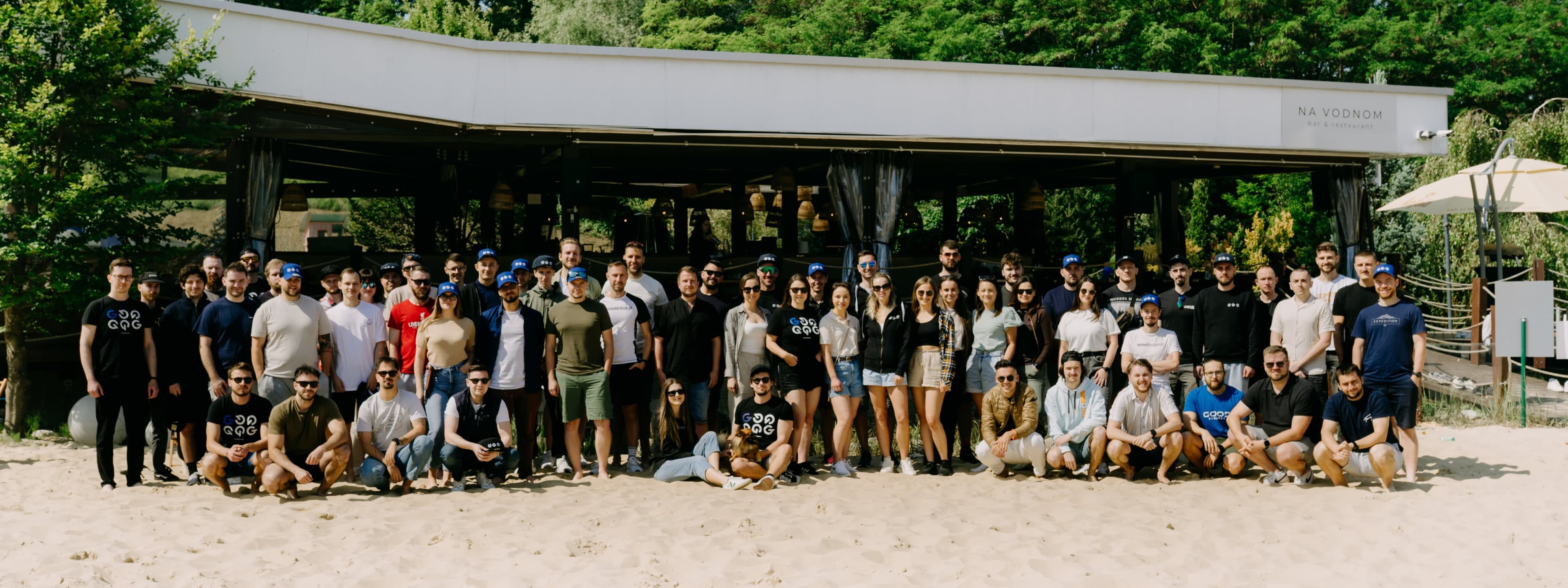 Veľká skupina ľudí pózuje na piesku počas firemného podujatia pod holým nebom. V pozadí sú stromy, prístrešok a modrá obloha.