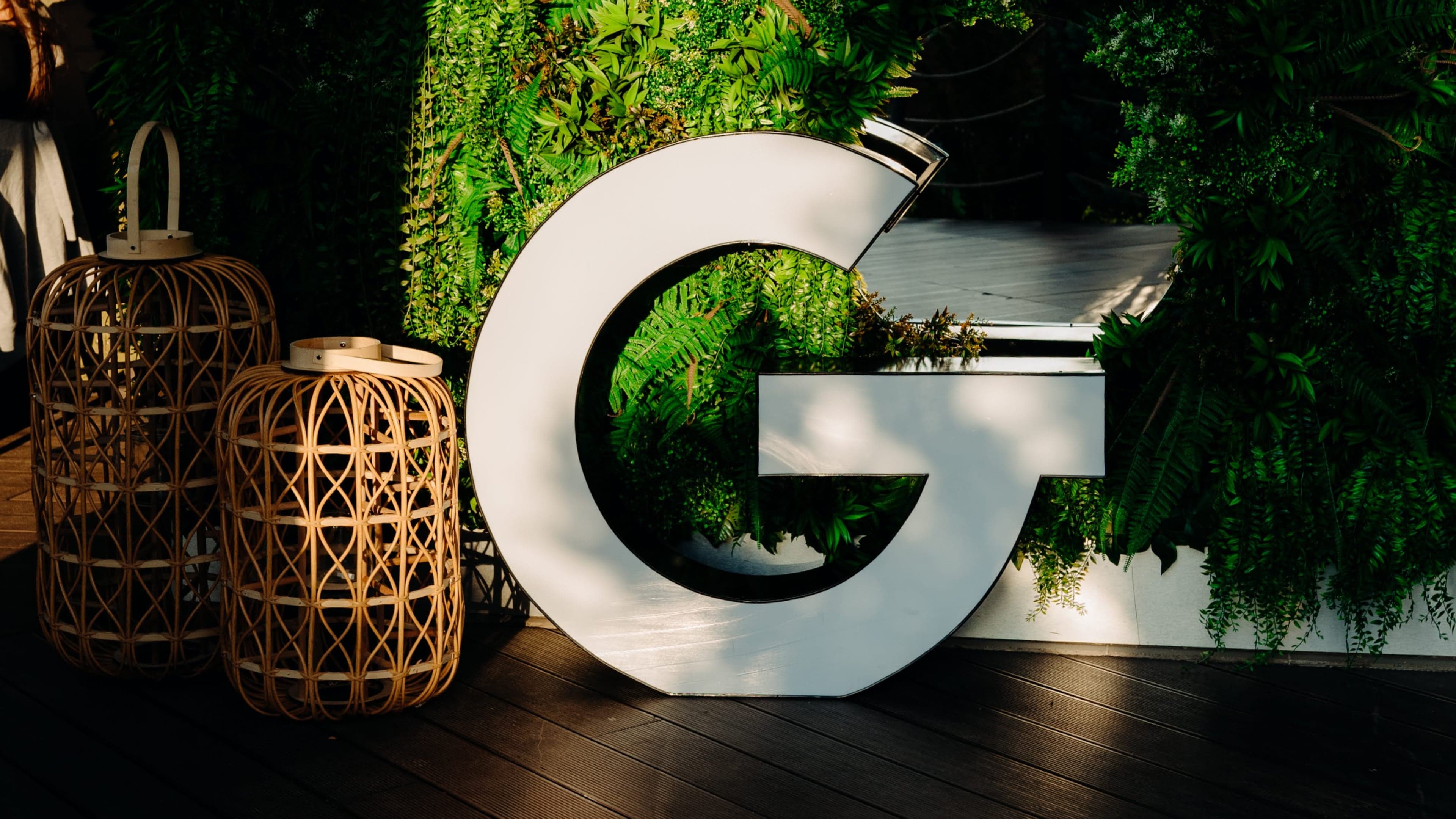 Biele logo spoločnosti GoodRequest postavené pred stenou pokrytou rastlinami, vedľa sú drevené lampáše.