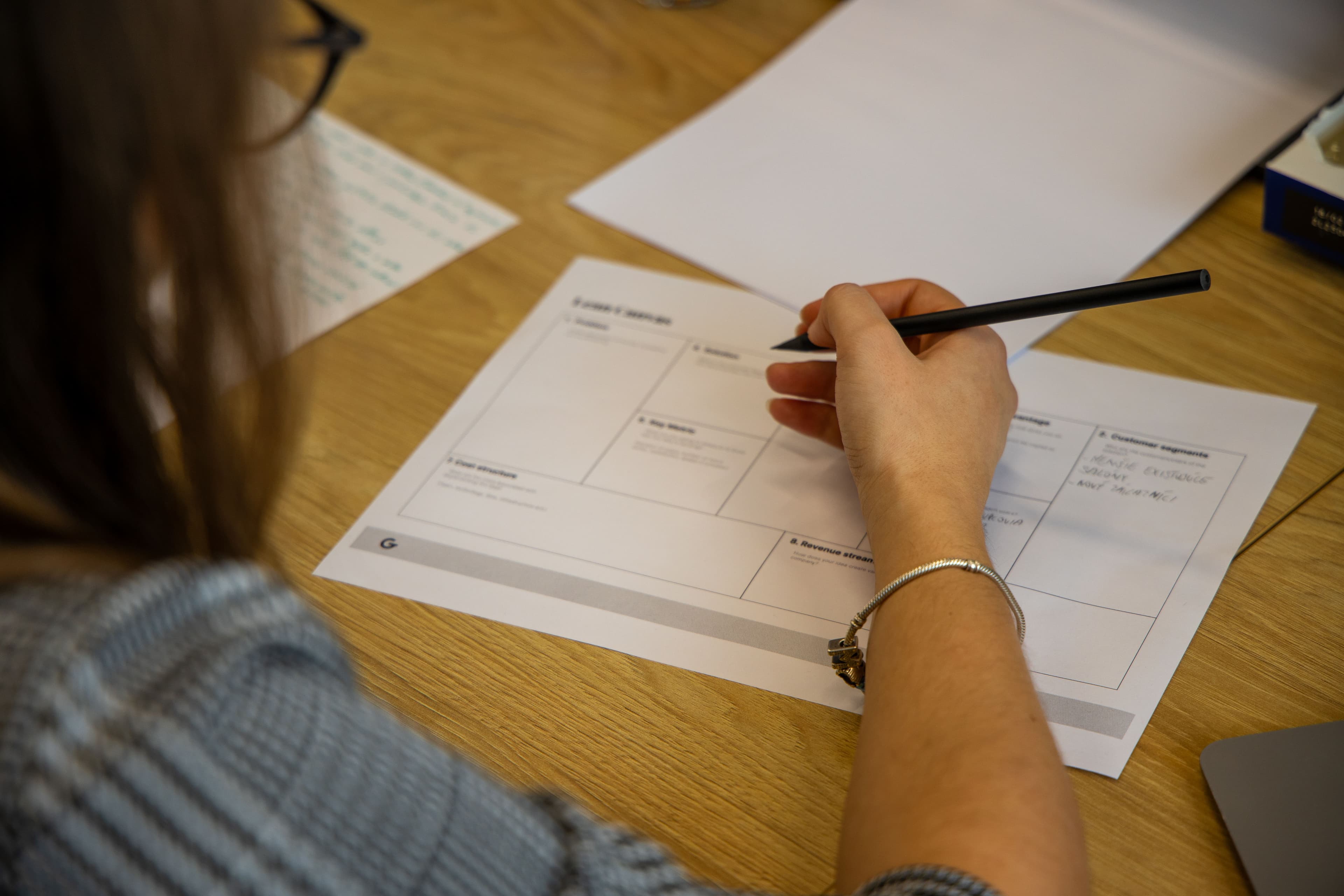 Osoba píše čiernym perom do pracovného listu na drevenom stole. Na papieri sú sekcie s nadpismi a riadkami na vyplnenie. V pozadí je ďalší papier s ručne písanými poznámkami.